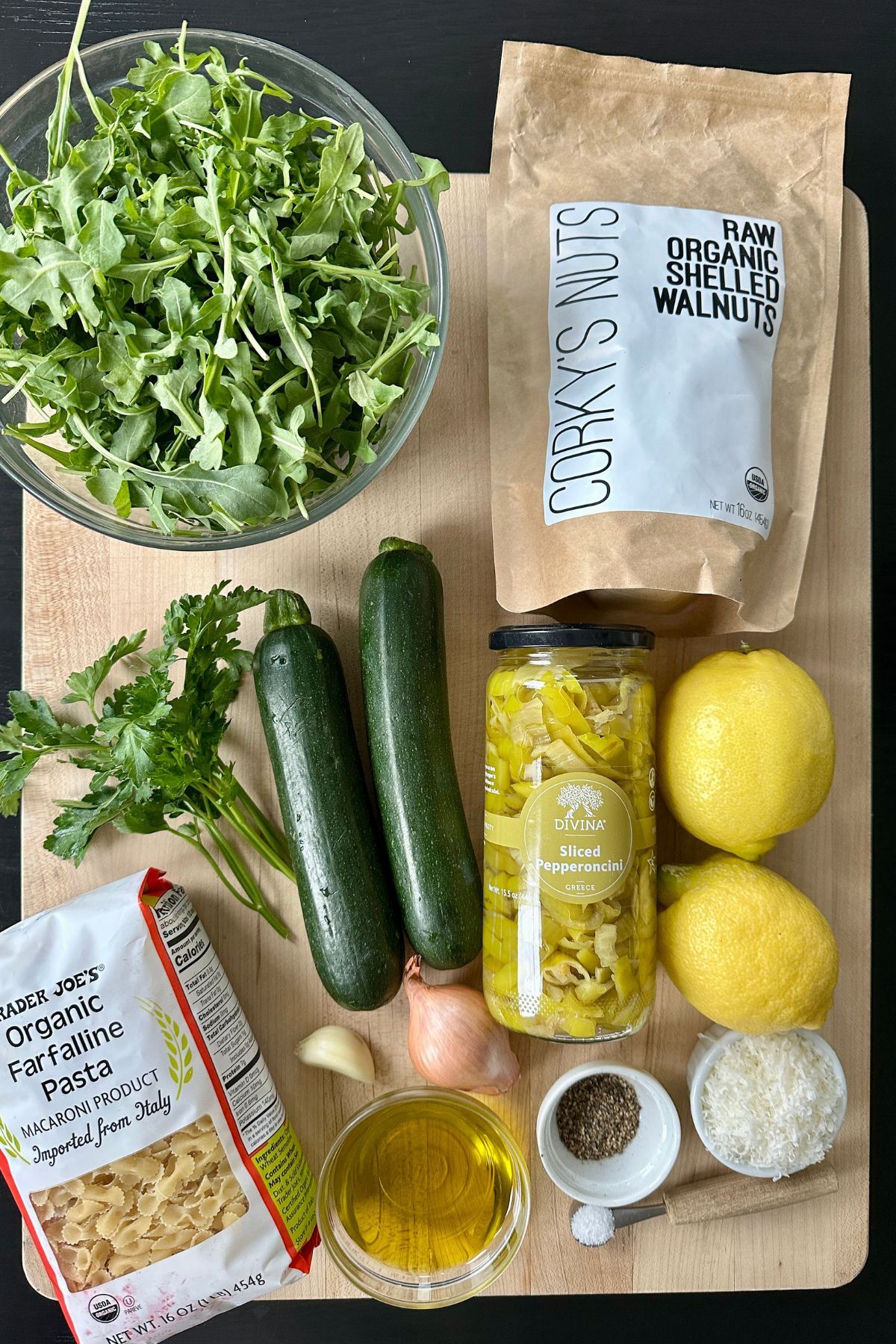 Ingredients for arugula pasta salad including arugula, zucchini, pasta, pepperoncini, walnuts, garlic, shallot, lemon, parsley, Parmesan, and olive oil arranged on a board
