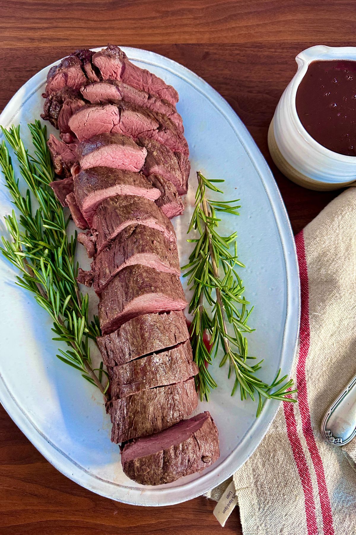 Beef tenderloin sliced into rounds and plated with rosemary sprigs beside it, with a gravy boat of red wine sauce next to the serving plate.