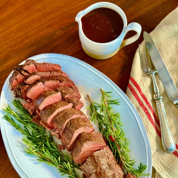 Beef tenderloin sliced and served on a white platter with sprigs of rosemary around it and a small gravy boat of red wine sauce beside it.
