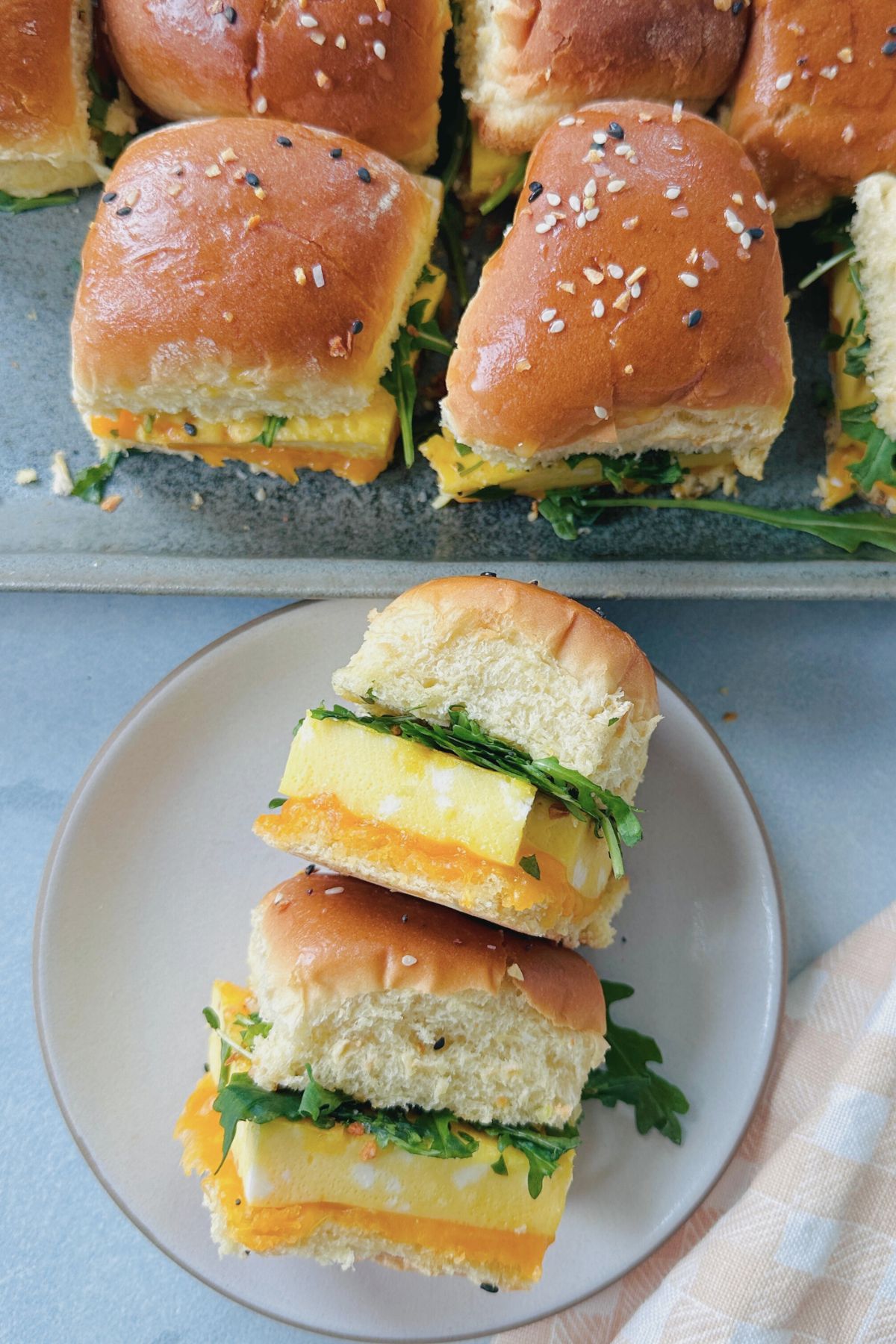 Breakfast slider on a plate, lying on its side to show the layers, with a tray of additional breakfast sliders in the background.