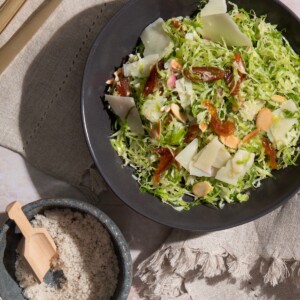 Black bowl of brussels sprouts salad with golden raisins, almonds, and shaved Parmesan in truffle vinaigrette, with a small bowl of sea salt, knife, and fork alongside.