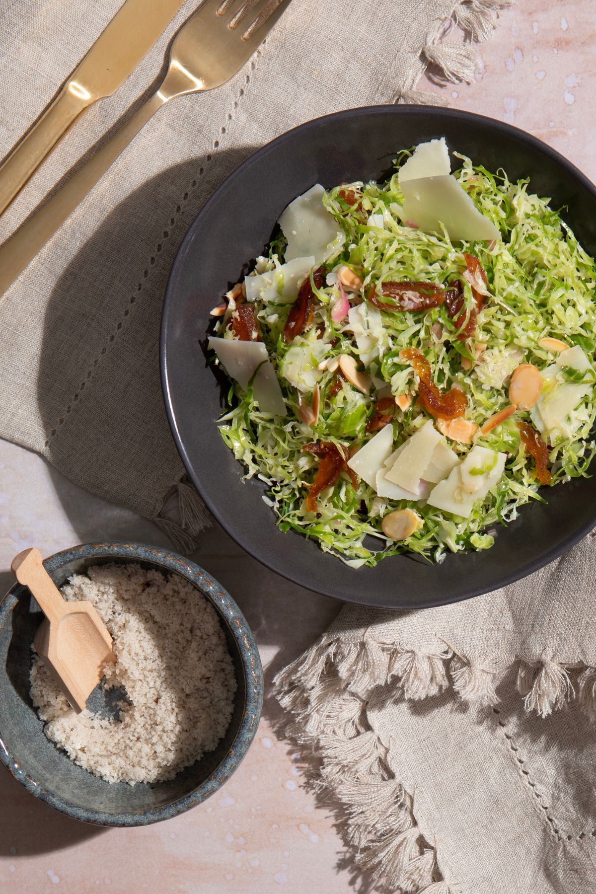 Black bowl of brussels sprouts salad with golden raisins, almonds, and shaved Parmesan in truffle vinaigrette, with a small bowl of sea salt, knife, and fork alongside.