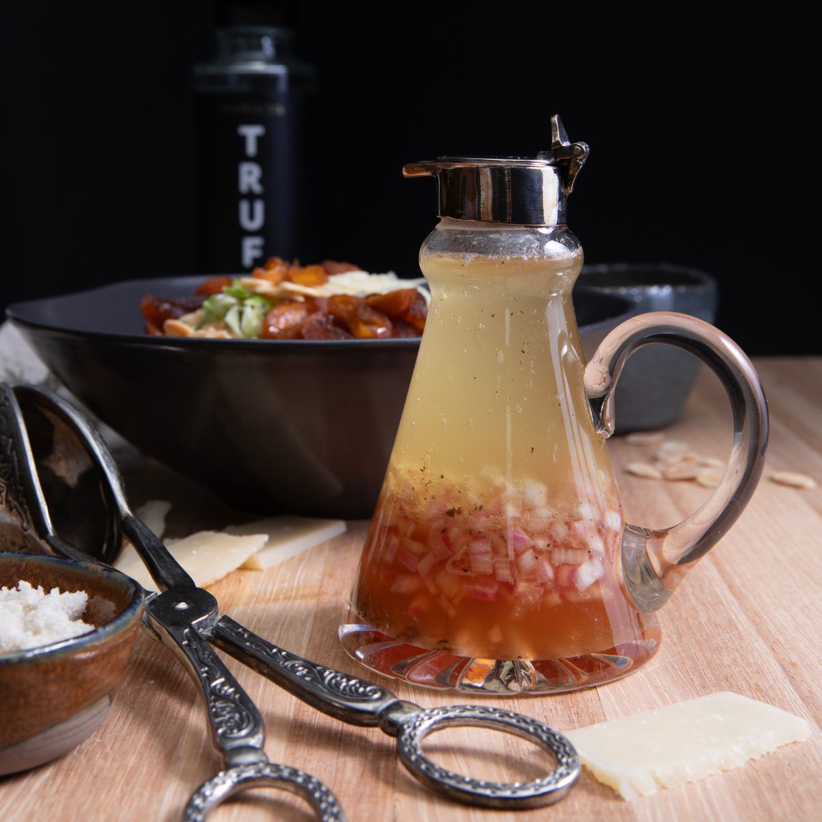 Glass jar of truffle oil vinaigrette on a light wooden counter with a black bowl of brussels sprouts salad in the background.