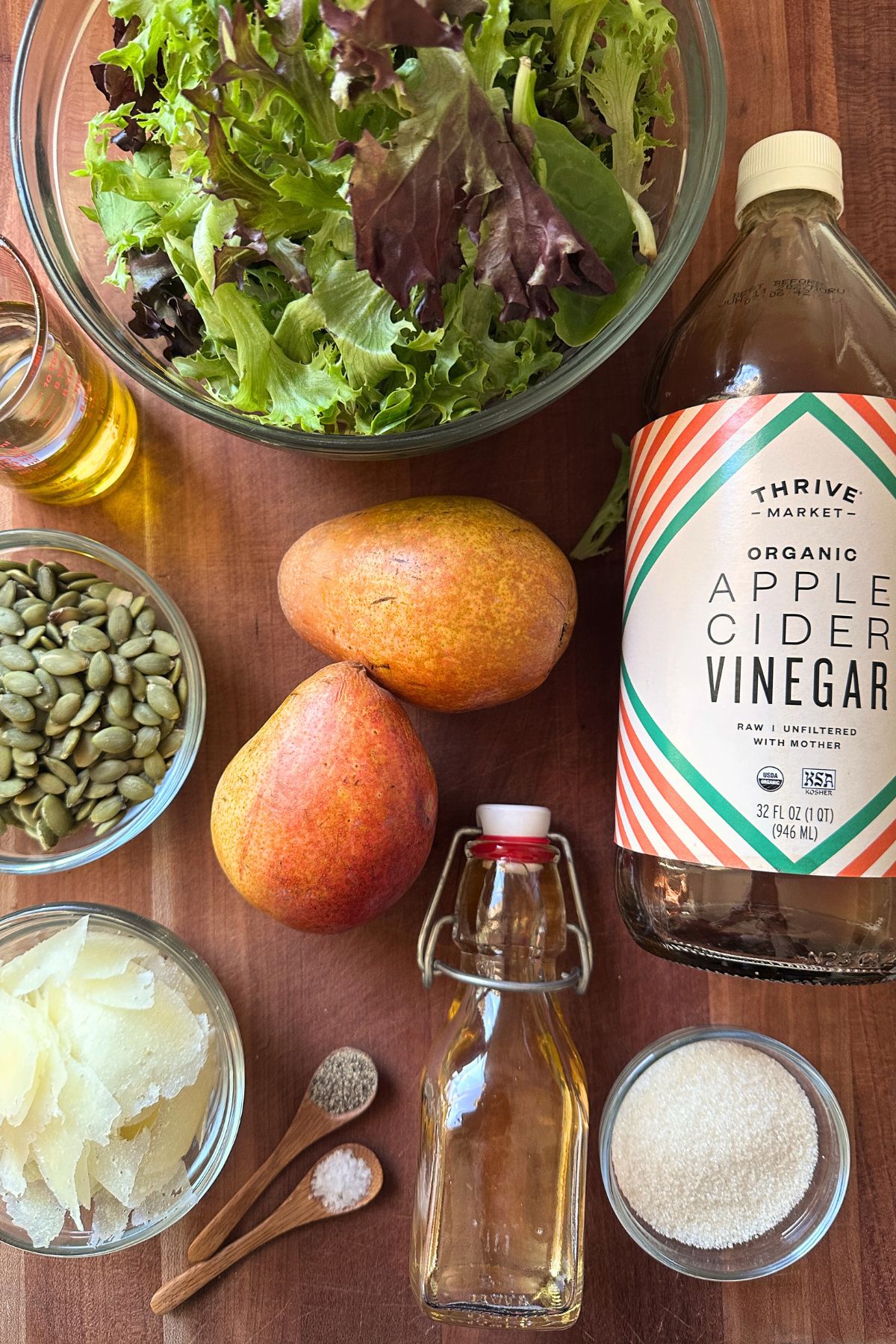 Ingredients for caramelized pear salad on a wooden counter top including greens, apple cider vinegar, pears, pumpkin seeds, cheese, maple syrup, oil and seasonings.