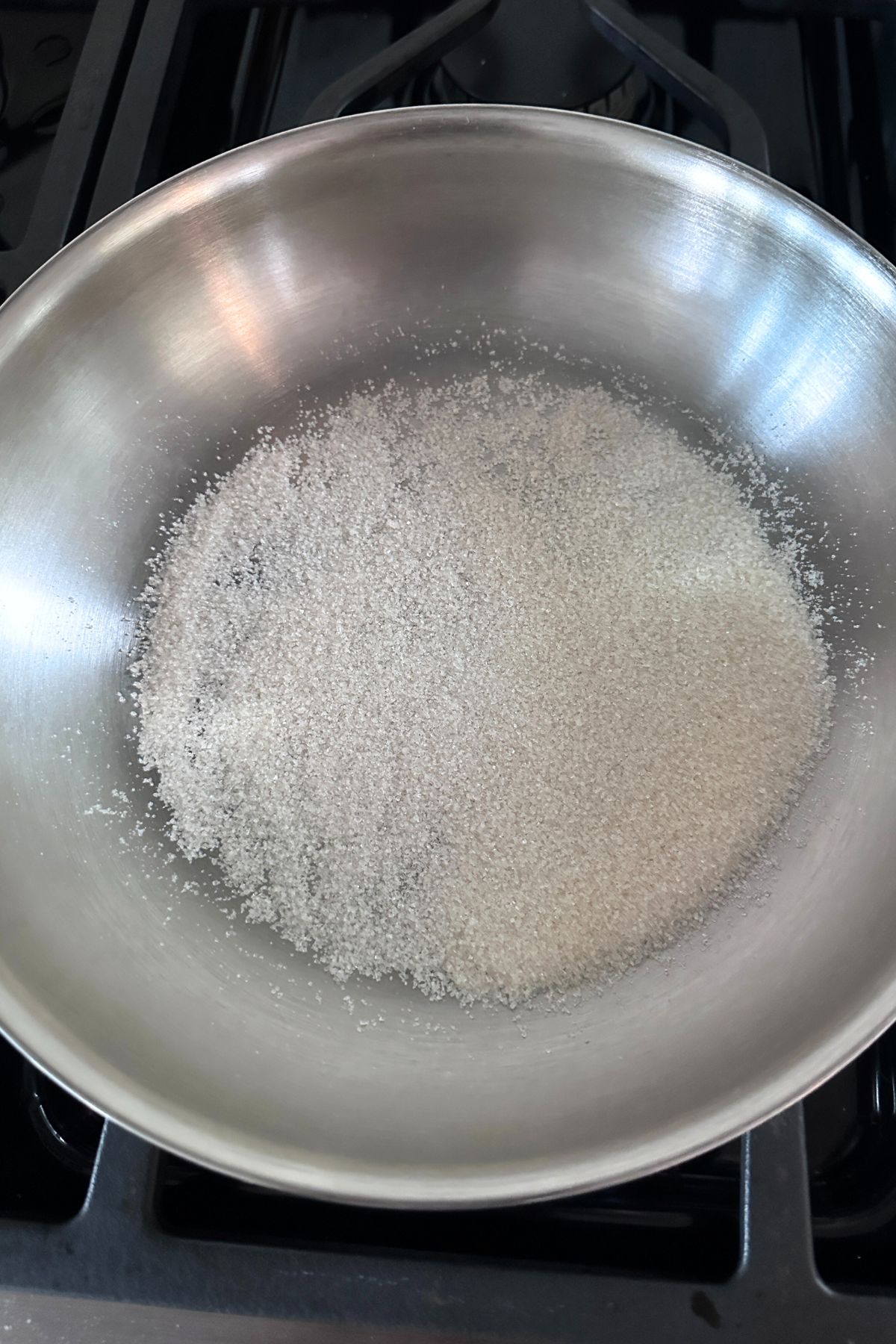 Silver skillet on a stovetop with sugar spread evenly, ready to melt for caramelizing.