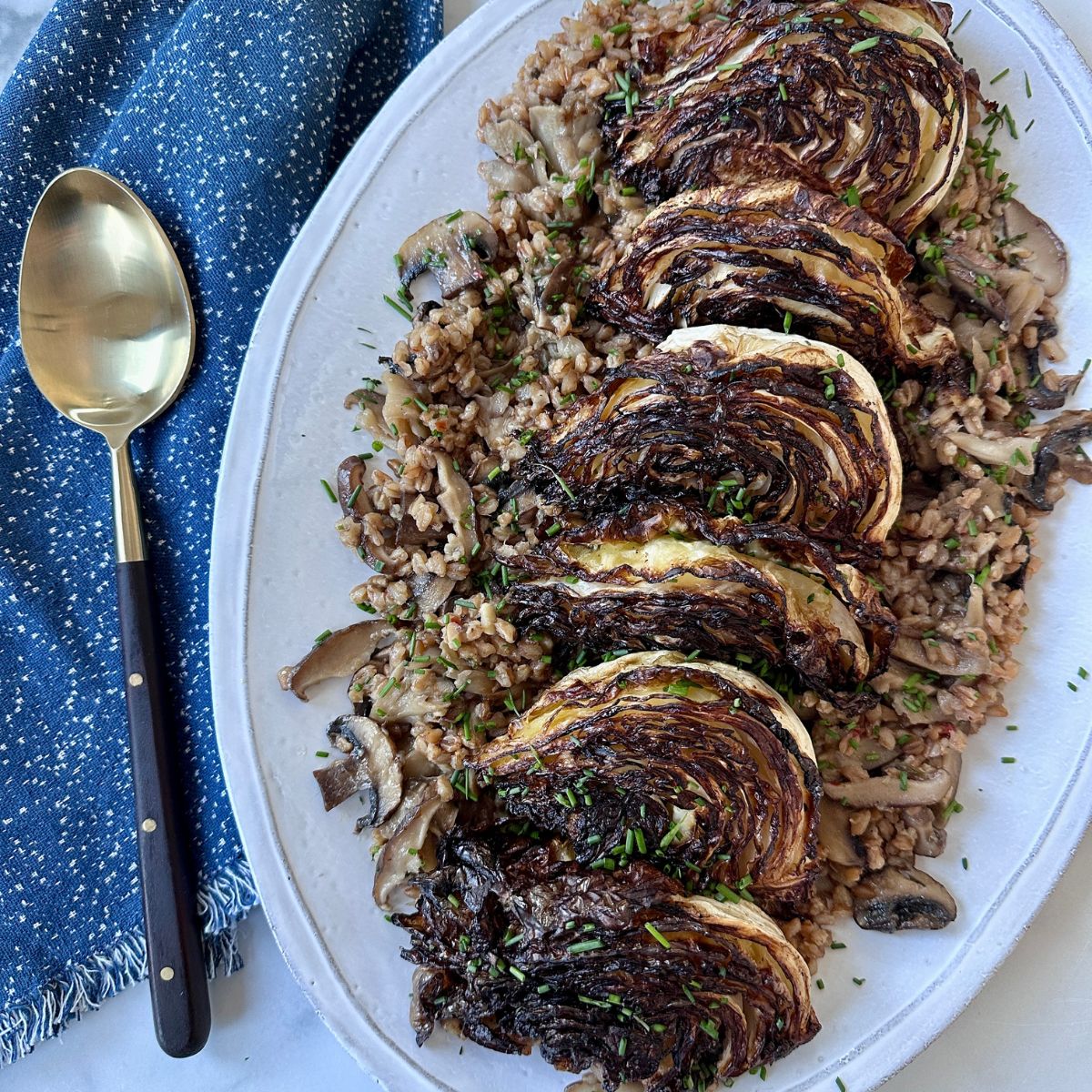 Mushroom farro topped with charred cabbage on a white serving platter, with a navy blue hand towel and a serving spoon beside it.