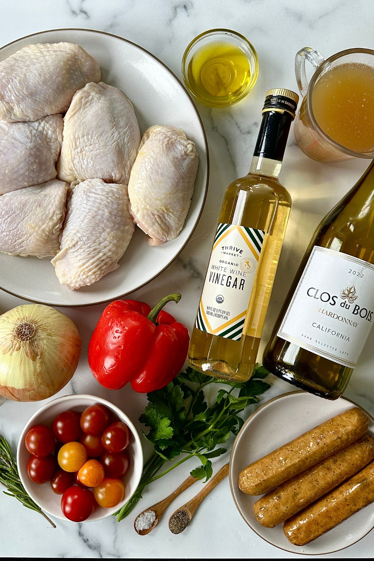 Overhead shot of raw ingredients for chicken scarpariello including chicken thighs, Italian sausage, bell peppers, onions, garlic, rosemary, and cherry tomatoes on a white counter.