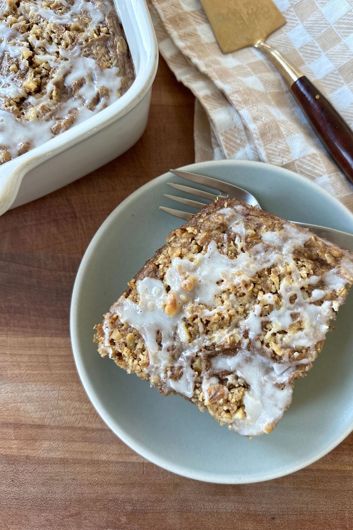 A slice of coffee cake baked oatmeal served on a blue plate with a fork beside it, with the remaining baked oatmeal visible in a white baking dish in the background.