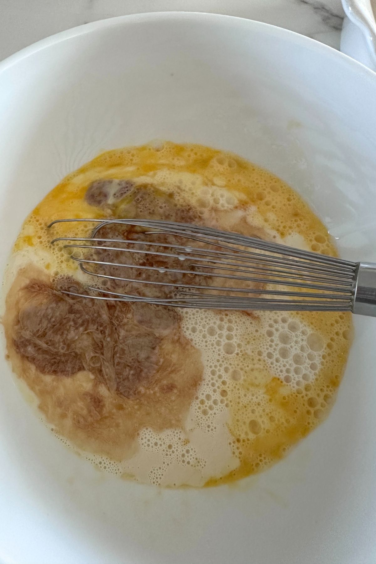 A large white bowl with eggs, milk, sweetener, and vanilla being whisked.