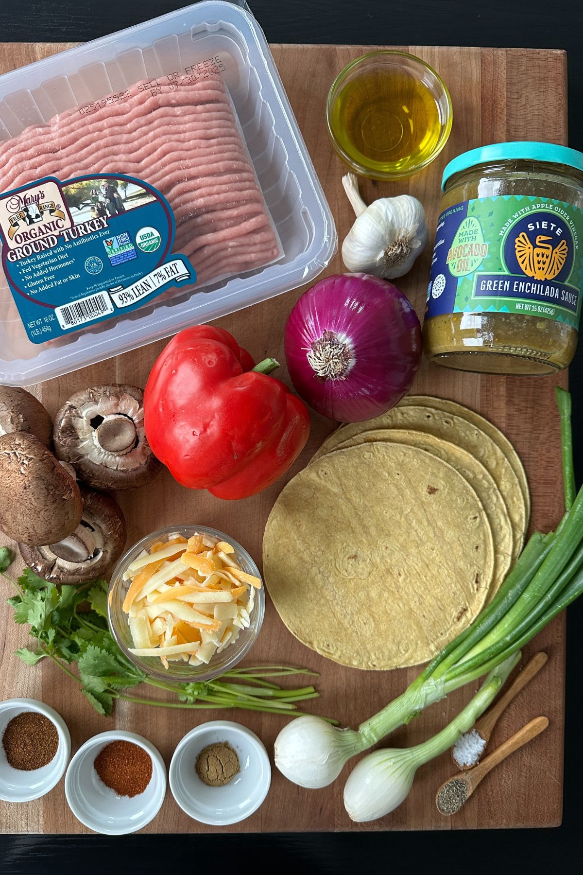 Overhead view of fresh green chile enchilada ingredients including tortillas, bell peppers, zucchini, onions, garlic, cheese, and cilantro on a white surface.