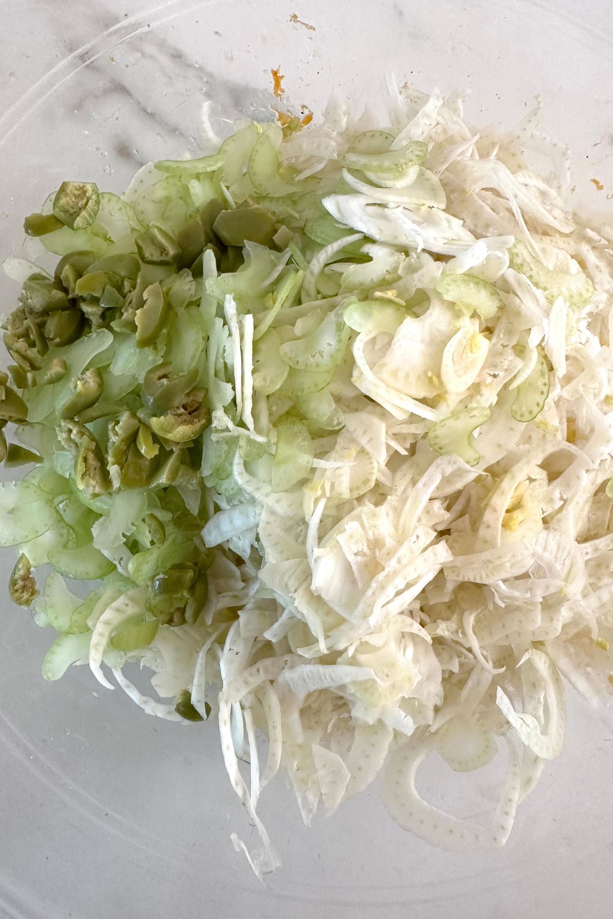 Sliced fennel stems mixed with chopped fennel fronds and green olives in a bowl, ready to be dressed for fennel salad.