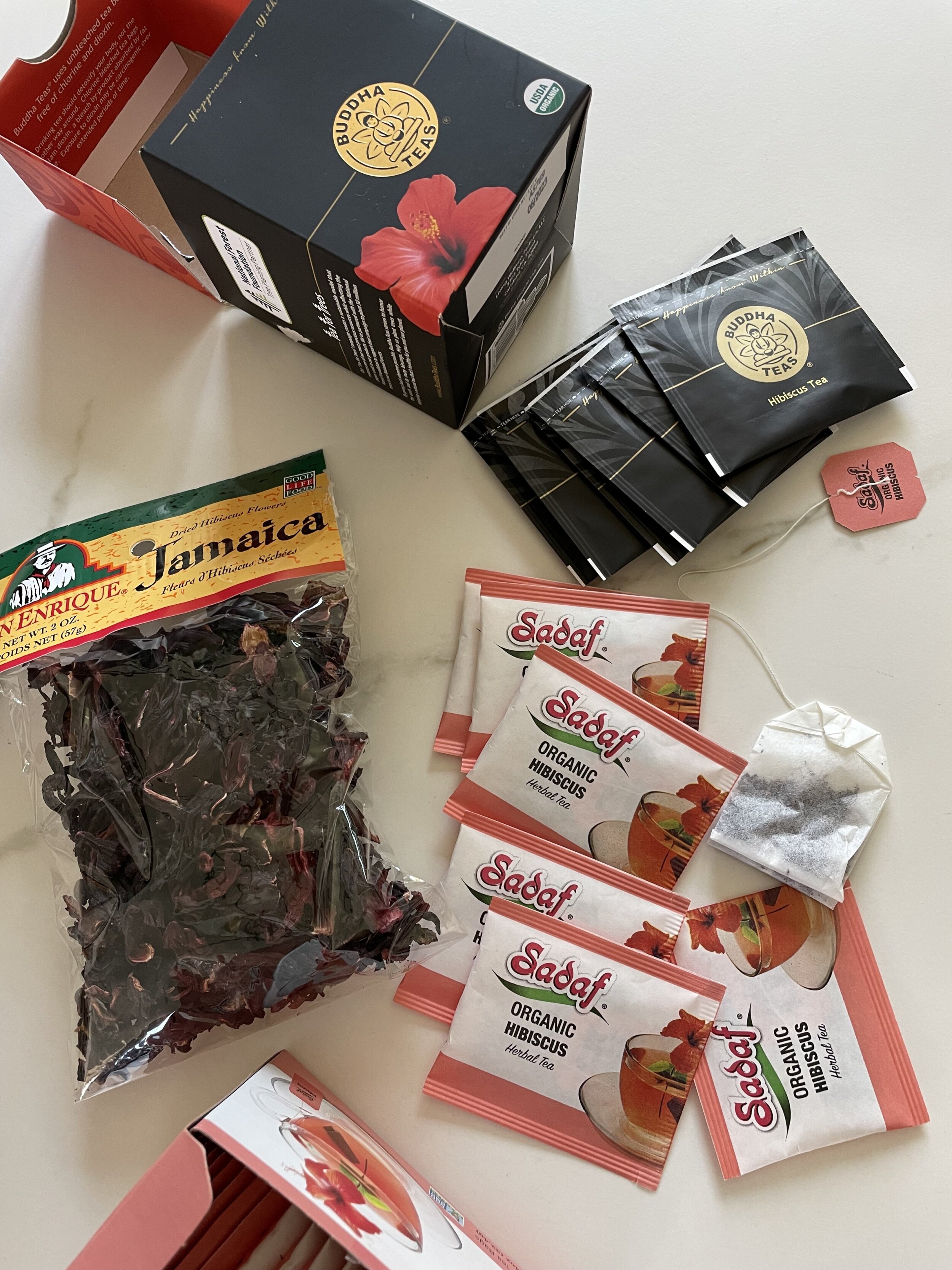 Different brands of hibiscus tea on a white countertop, including Buddha Teas, Sadaf Organic Hibiscus Herbal Tea, and dried Jamaica hibiscus flowers.