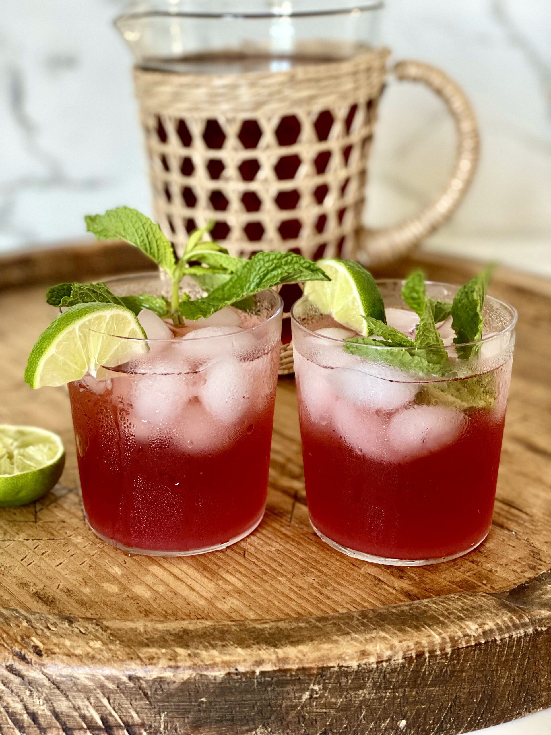 Two glasses of red hibiscus mocktails with ice, lime slices, and mint on a wooden cutting board, with a pitcher of hibicus mocktail in the background.
