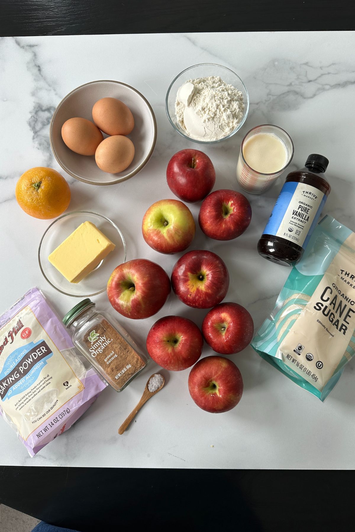 Recipe ingredients for invisible apple cake including Honeycrisp apples, butter, eggs, flour, cane sugar, orange, nutmeg, and vanilla on a white surface.