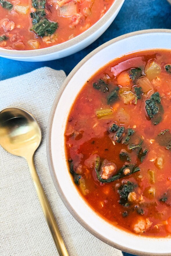 Two bowls of sausage lentil soup on a table, with a linen napkin and a gold spoon beside the main bowl.