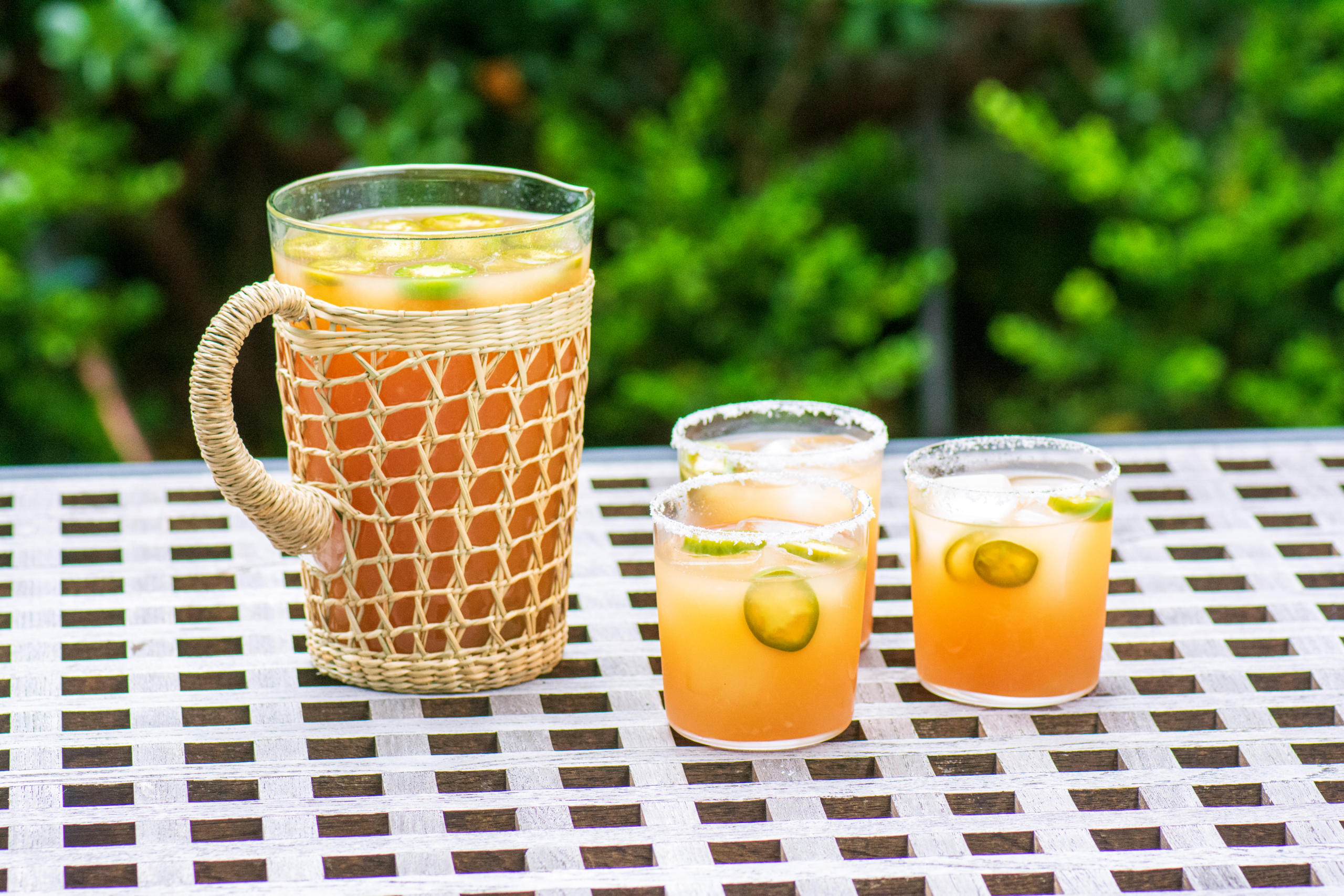 Pitcher of spicy margaritas with three salt-rimmed glasses over ice with jalapeno slices on an outdoor table.