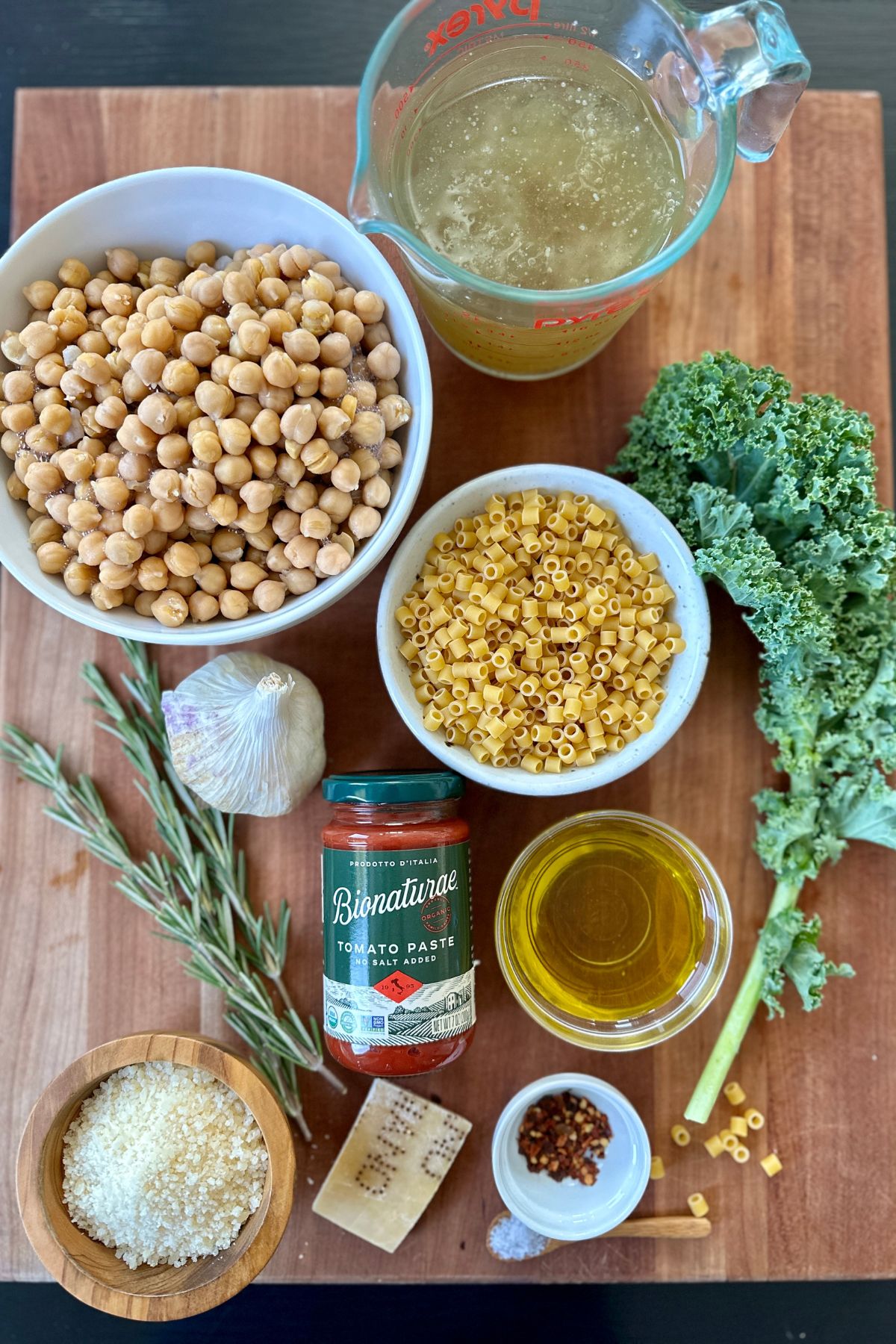Ingredients for Pasta e Ceci including olive oil, garlic, rosemary, tomato paste, chickpeas, Parmesan rind, broth, pasta, and Parmesan cheese on a wooden cutting board.