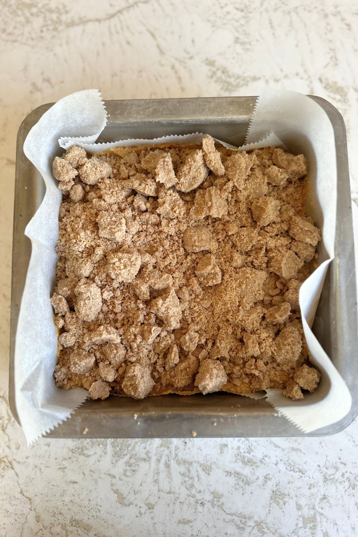 Pumpkin coffee cake batter in an 8-inch parchment-lined pan topped evenly with chilled cinnamon crumb mixture.