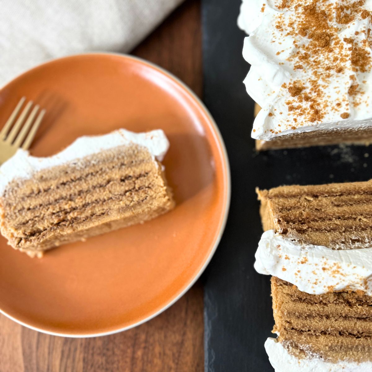 A slice of pumpkin icebox cake on an orange plate next to a larger partially sliced pumpkin icebox cake on a black serving platter.