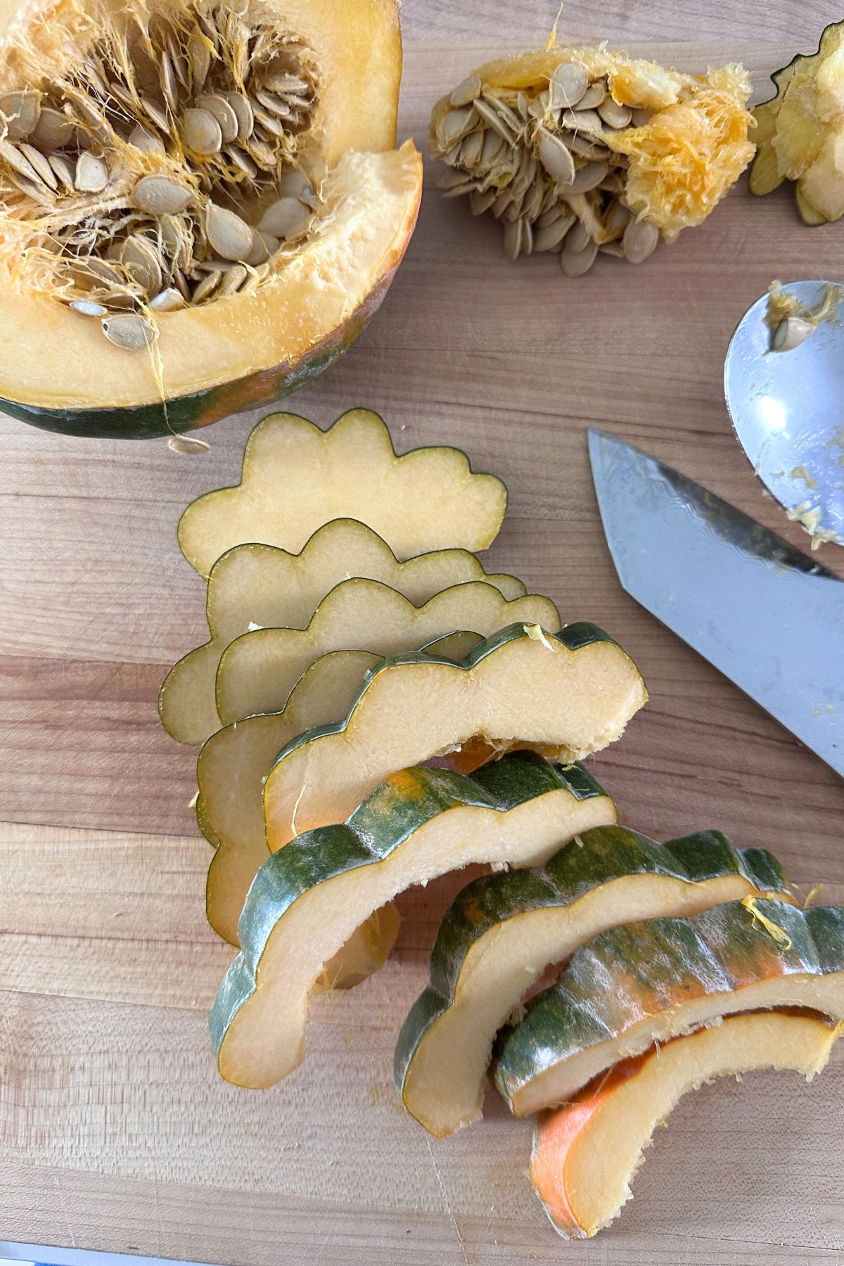 Halved acorn squash with seeds exposed and some scooped out and placed beside it, with several scalloped-cut pieces arranged on a light wooden cutting board.