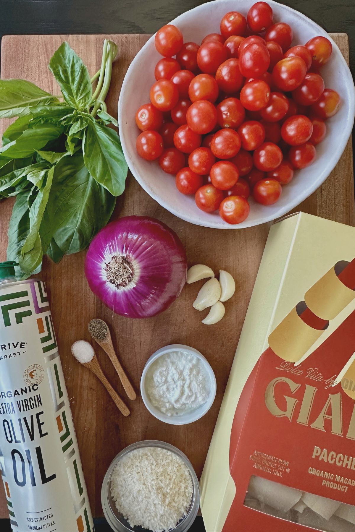 recipe ingredients on a wooden cutting board including fresh cherry tomatoes, red onion, ricotta cheese, olive oil, fresh garlic and basil. 