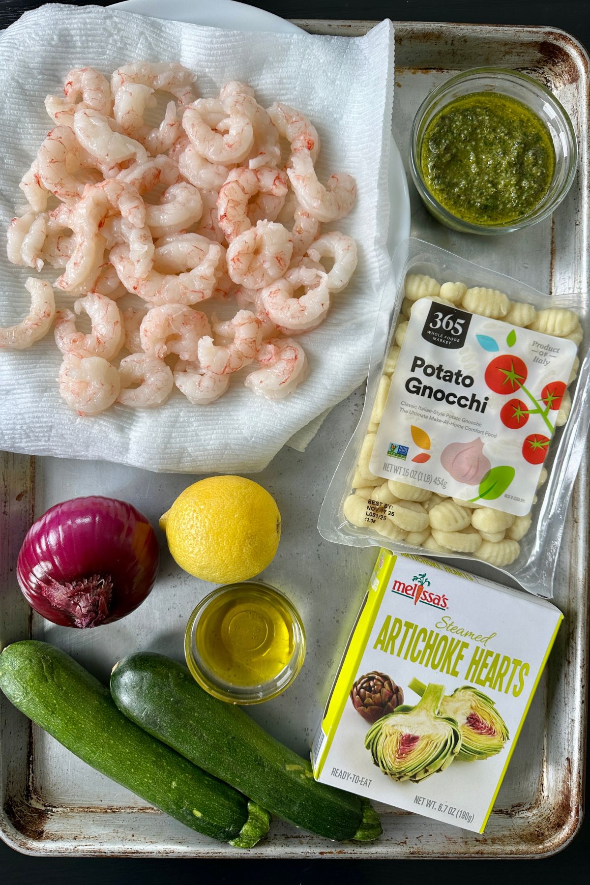 Ingredients for sheet pan gnocchi laying on a baking sheet including uncooked shrimp, pesto, potato gnocchi, red onion, lemon, olive oil, steamed artichoke hearts, and 2 zucchini. 