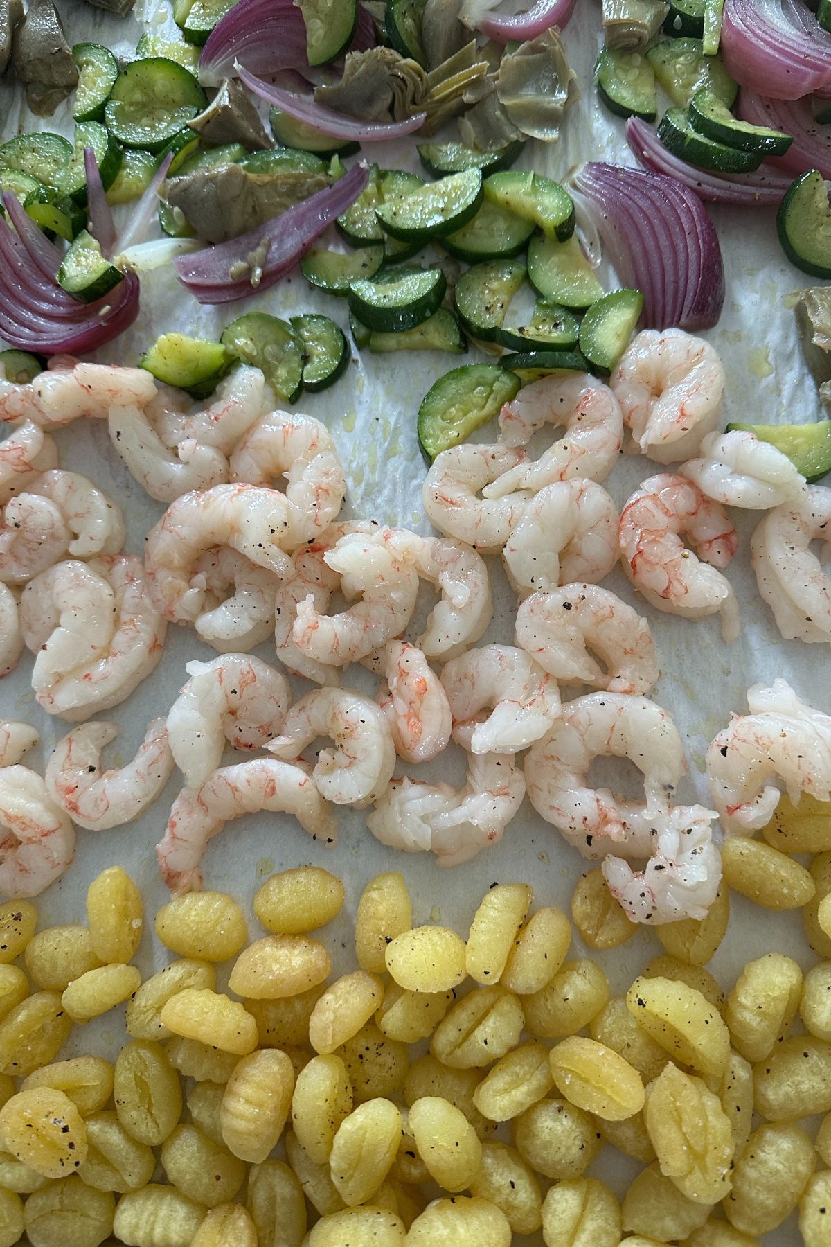 Uncooked shrimp arranged on a sheet pan alongside golden gnocchi and roasted vegetables before the final bake.