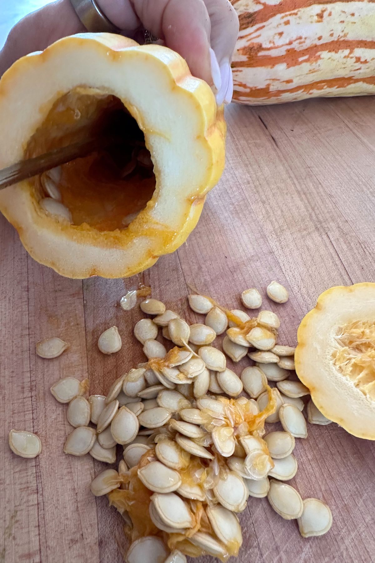 A spoon scooping seeds from a halved delicata squash, with the seeds resting on a wooden countertop.