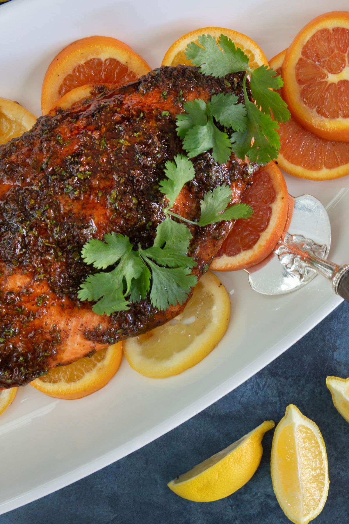 A glazed salmon fillet garnished with cilantro is served on a white platter, surrounded by orange and lemon slices. A silver serving utensil is placed on the right side. The background has more lemon wedges on a dark surface.
