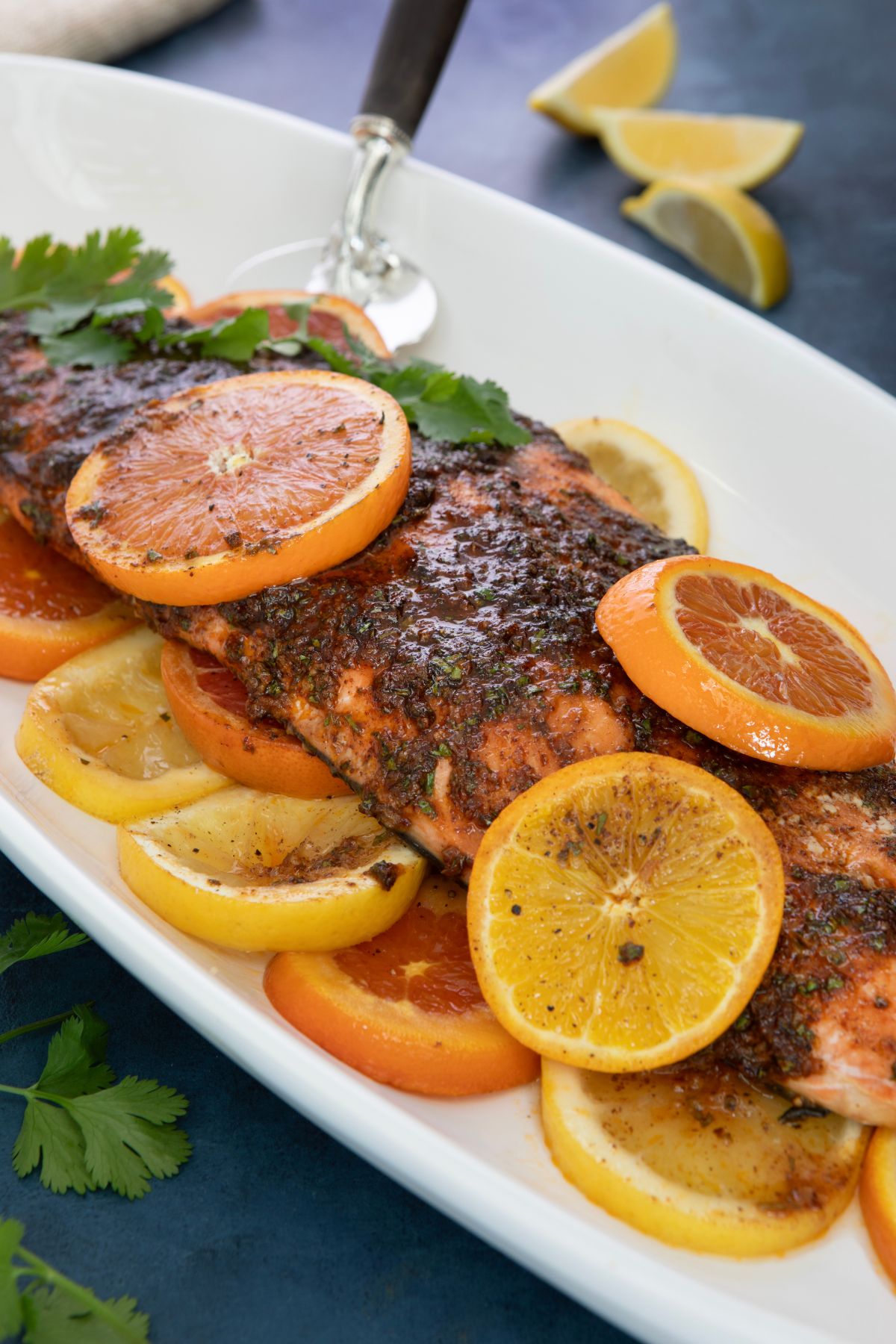 A cooked salmon fillet topped with herbs and spices is garnished with orange slices. The fish is served on a white platter, surrounded by lemon and orange slices. Fresh cilantro is placed on top, and lemon wedges are in the background.