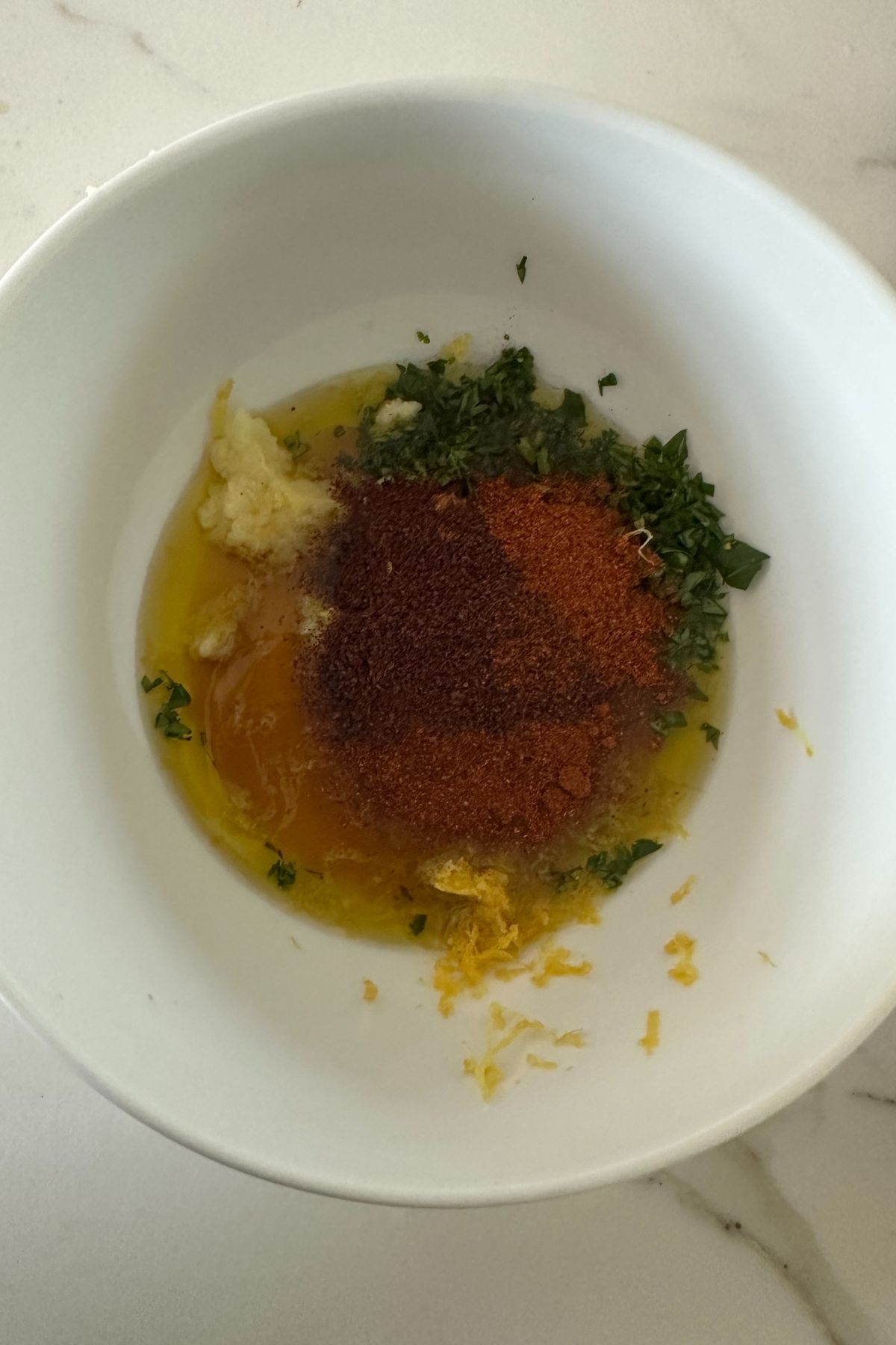 A white bowl contains minced garlic, chopped herbs, ground spices, yellow zest, and olive oil. The ingredients are partially mixed, sitting on a white countertop.