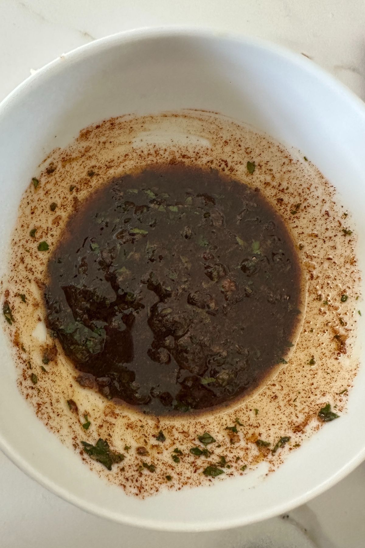 A white bowl contains a dark brown liquid with visible herbs and spices. The mixture appears to be a marinade or sauce, with finely chopped green herbs dispersed throughout. The bowl sits on a white surface.