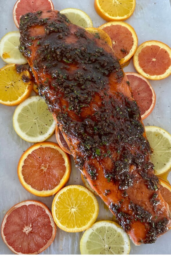 Grilled salmon fillet topped with a dark herb glaze, surrounded by slices of orange, lemon, and grapefruit on a white surface.