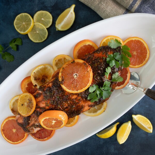 A cooked salmon fillet garnished with slices of lemon and orange is displayed on a white oval platter. Fresh cilantro is placed on top. Lemon and orange slices are scattered around the dish on a dark blue background.