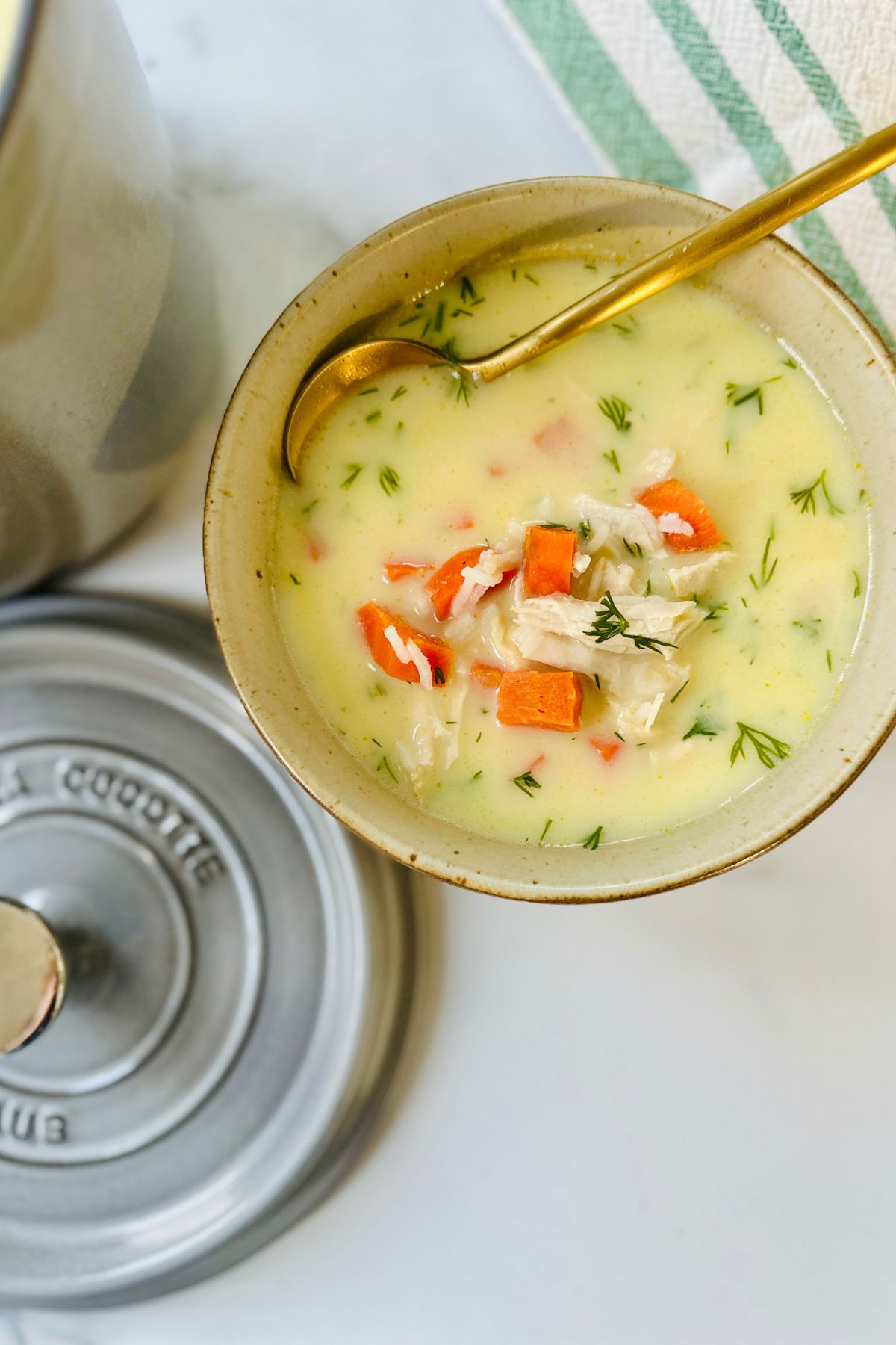 A small bowl of avgolemono soup with leftover turkey and carrots, a spoon resting inside, on a white countertop beside a green-and-white striped towel and the lid of a Dutch oven.
