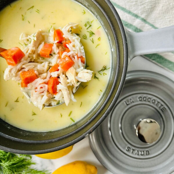 A large gray Dutch oven filled with avgolemono soup, with a ladle lifting a scoop that shows the leftover turkey, carrots, and cooked rice.